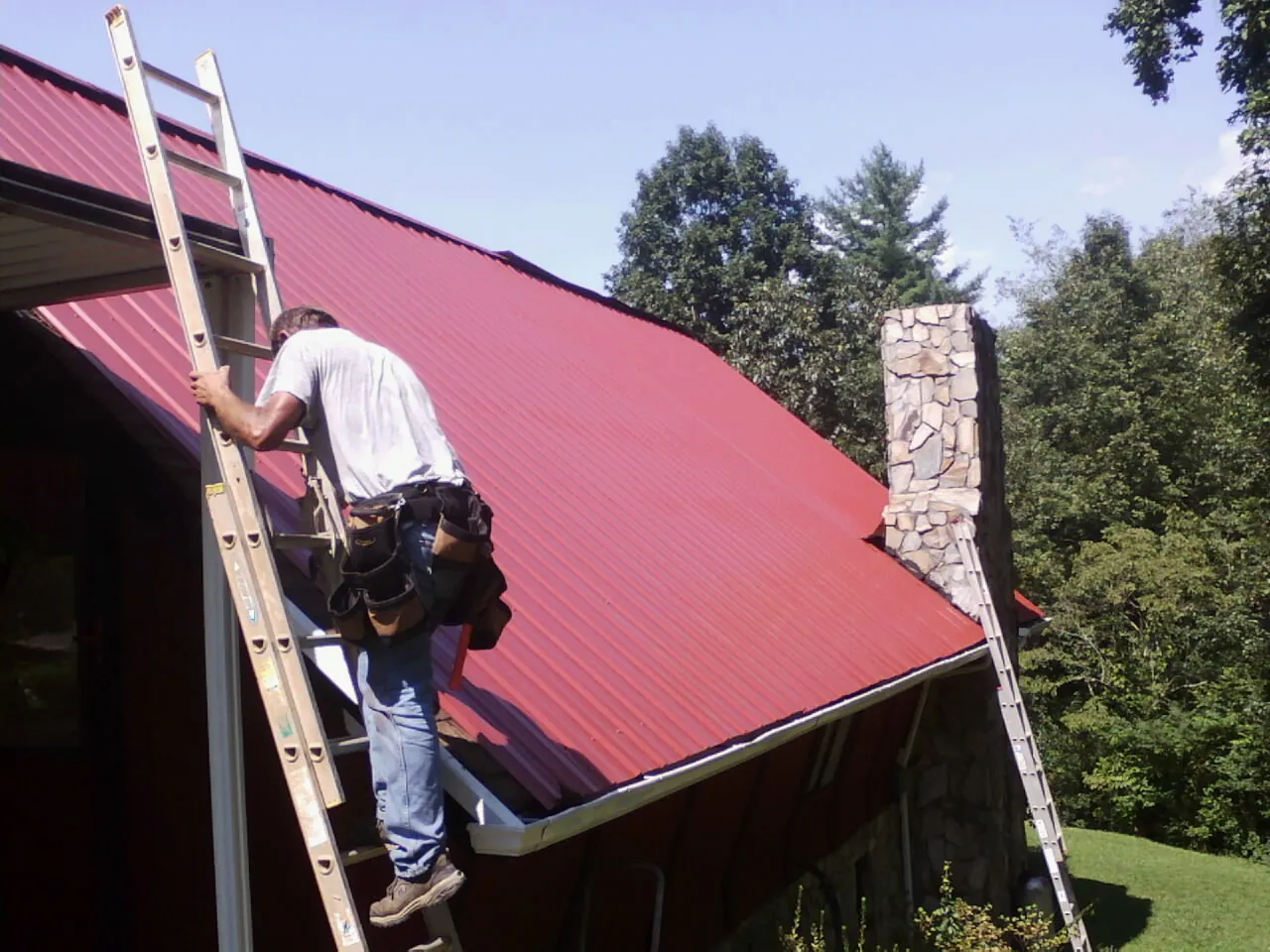 Roofer on a ladder beside a red metal roof and stone chimney — Soffit & Fascia Repair by Roofing Brothers in Galveston